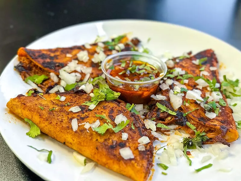 Image: Three Quesabirria tacos topped with raw onions and cilantro with birria dipping sauce, available at Fireside Chicken and Taco's newest location in Dallas.