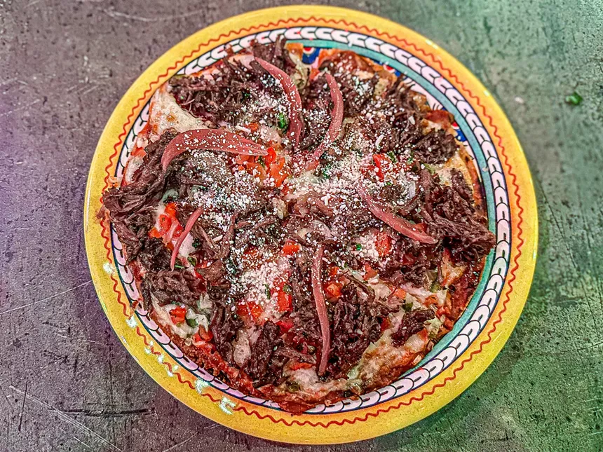 The Ranchero Pizza is a flour tortilla with a refried beans base, topped with spicy red salsa, cheese, barbacoa, pico de gallo, pickled red onions, peppers and queso fresco.